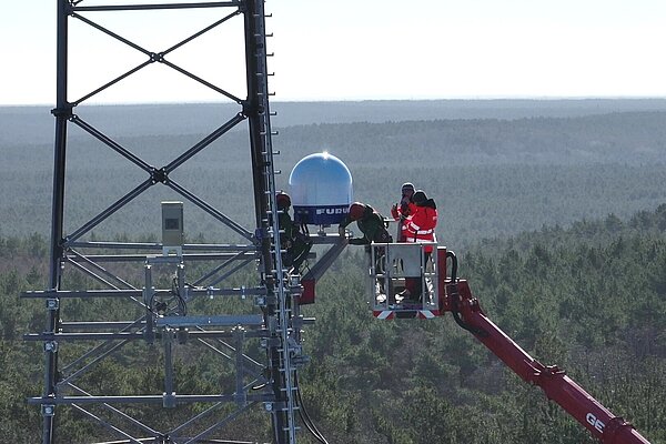 People are standing in a crane, installing a weather radar on a radio mast above the treetops.