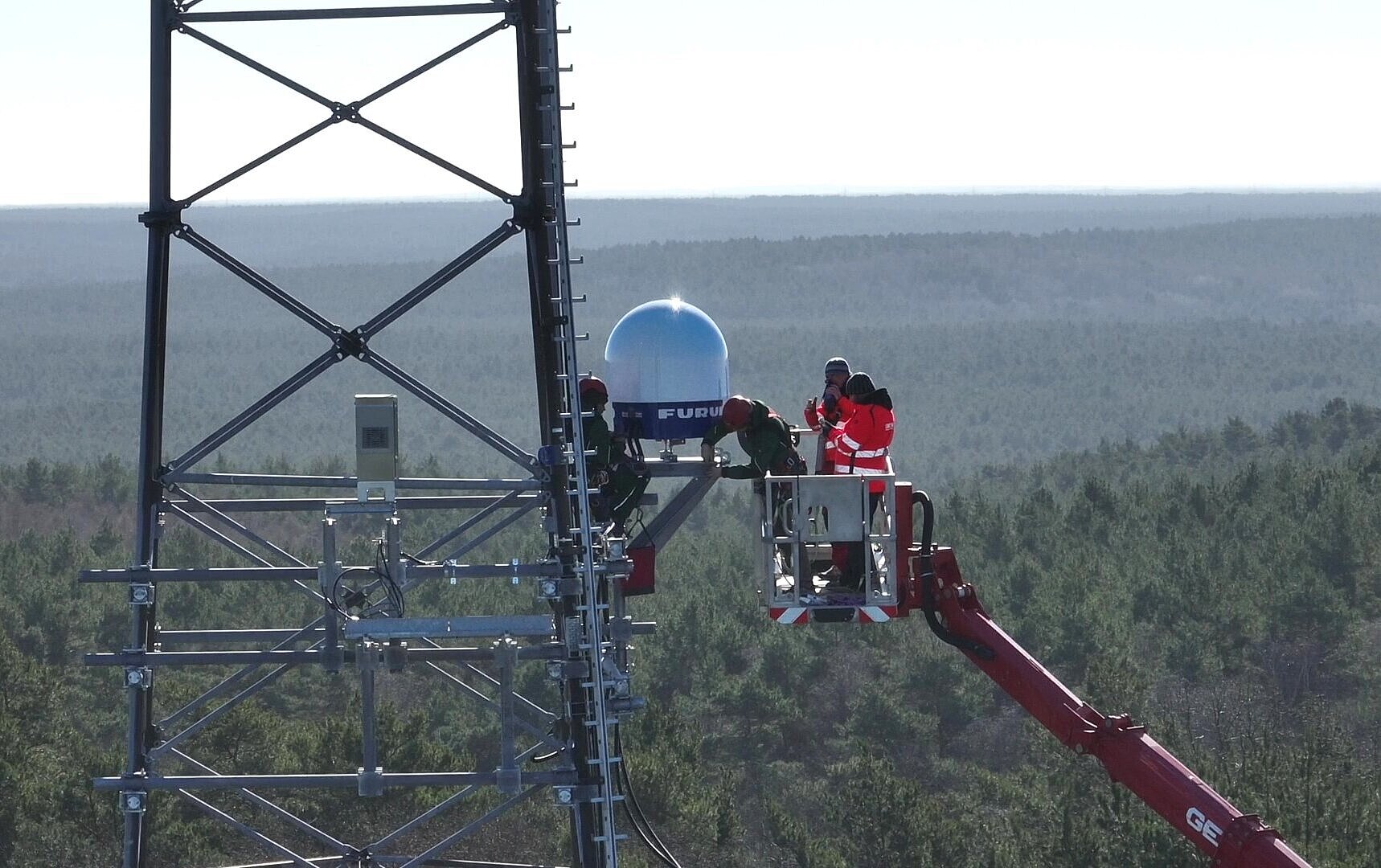People are standing in a crane, installing a weather radar on a radio mast above the treetops.