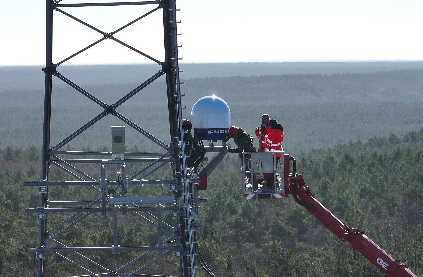 Menschen stehen in einem Kran und installieren an einem Funkmast über den Baumwipfeln einen Wetterradar.