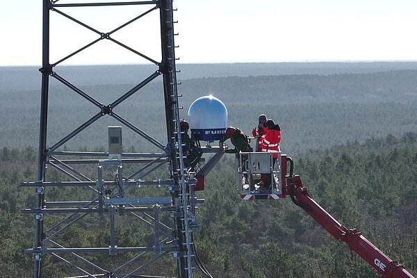 Menschen stehen in einem Kran und installieren an einem Funkmast über den Baumwipfeln einen Wetterradar.