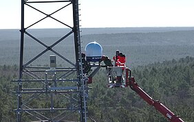 People are standing in a crane, installing a weather radar on a radio mast above the treetops.