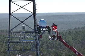 Menschen stehen in einem Kran und installieren an einem Funkmast über den Baumwipfeln einen Wetterradar.