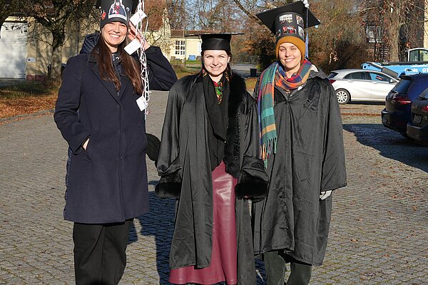 Drei Promovenden mit traditionellem Umhang und dekoriertem Doktorhut.