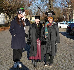 Drei Promovenden mit traditionellem Umhang und dekoriertem Doktorhut.